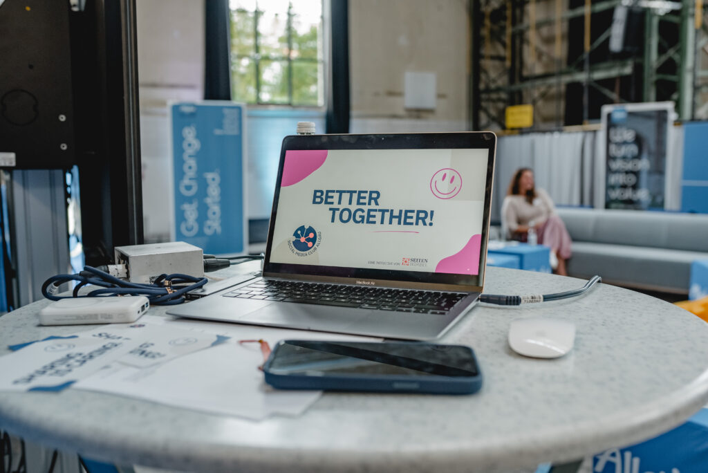 Zu Sehen ist ein Laptop auf einem runden Stehtisch in einem Veranstaltungsraum. Der Laptop ist geÃ¶ffnet und auf dem Bildschirm steht 